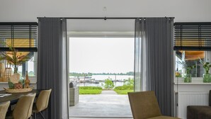 A modern living room with a large sliding glass door leading to a balcony with a view of a lake and boats. There are two chairs and a table with decorative items.