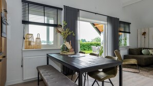 A modern kitchen with a dark countertop, a bench, and a dining table with chairs. There are windows with blinds, a radiator, and a view of greenery outside.