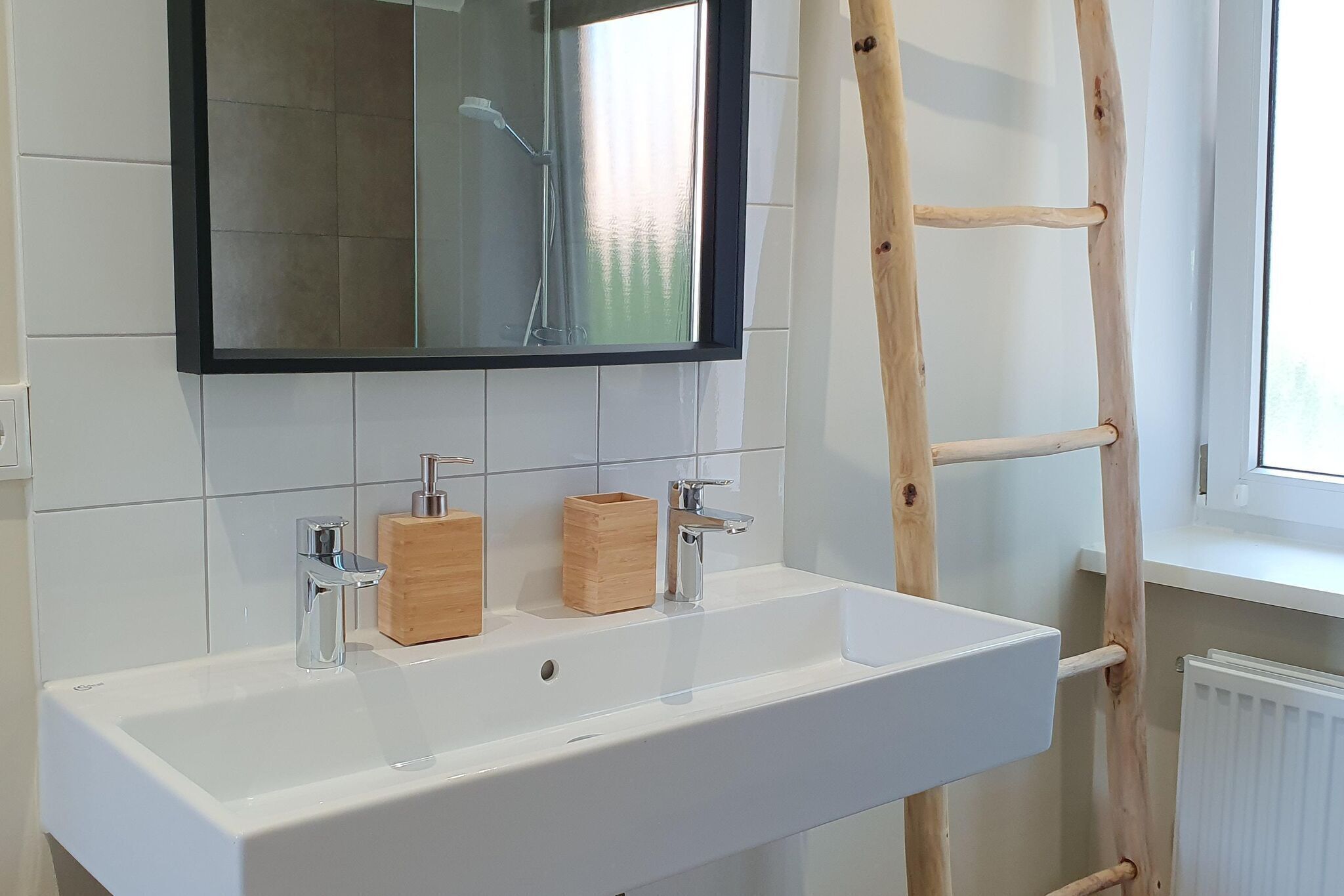 Une salle de bain avec un lavabo blanc, deux distributeurs de savon en bois, un grand miroir et une échelle en bois appuyée contre le mur.