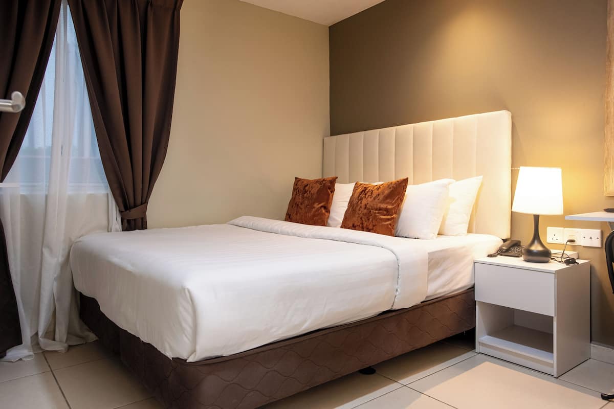 Una cama bien tendida con ropa blanca y dos almohadas marrones, una mesita de noche con lámpara, y un enchufe en la pared.