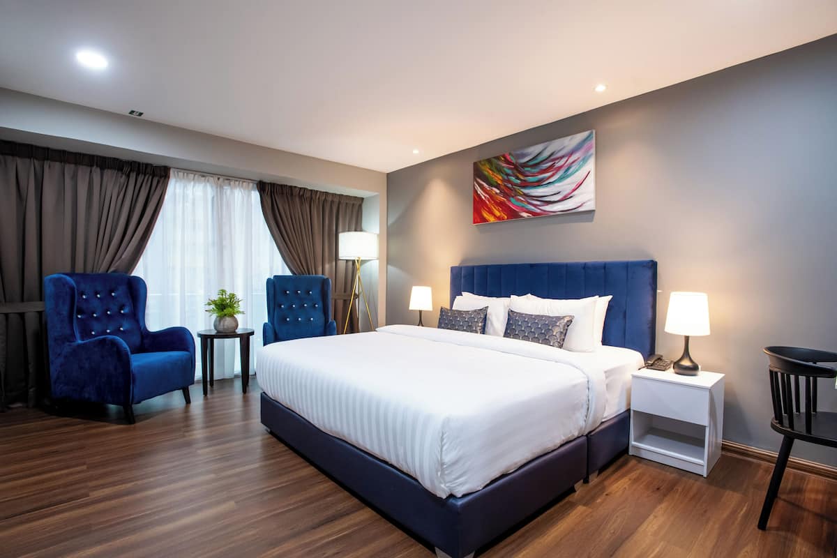 Una habitación de hotel moderna con una cama grande, dos sillones azules, una mesita de noche con lámpara y un elemento decorativo en la pared.