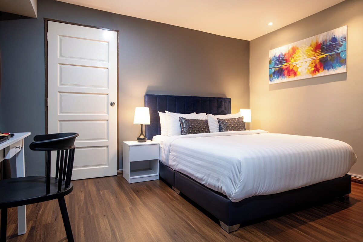 Quarto de hotel com cama, mesas de cabeceira, uma cadeira, escrivaninha e um quadro na parede.
