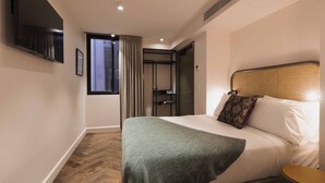 A hotel room with a bed, a small table, a TV mounted on the wall, and a window with curtains. - The Sarah by Urban Rest (Darlinghurst)