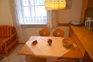 A cozy room with a wooden table, two heart-shaped chairs, a small table lamp, a radiator, and a window with curtains.