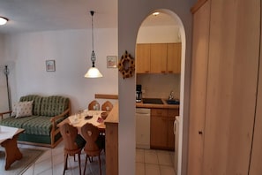 A living room with a sofa, dining table, and chairs, a kitchen with wooden cabinets and a dishwasher, and a hallway leading to another room.