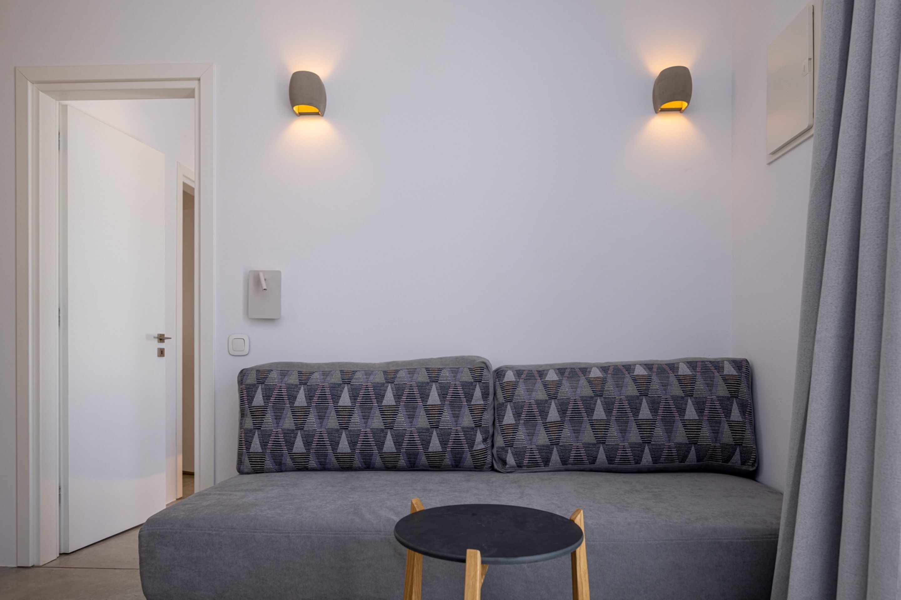Una habitación moderna con un sofá gris, dos cojines estampados, una mesita redonda y dos luces fijadas a la pared.