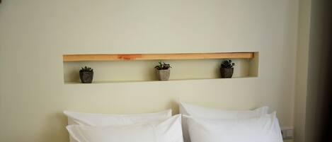 A bed with white bedding, a wooden nightstand, and a shelf with three potted plants.