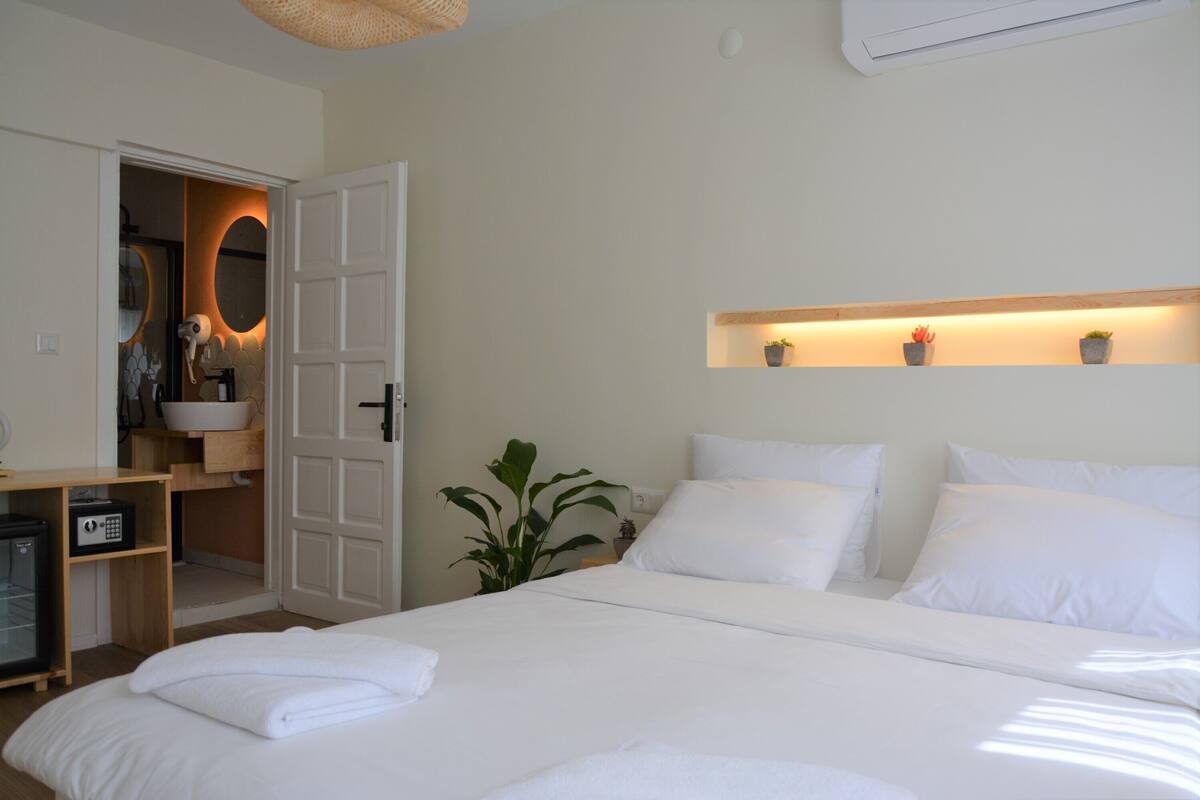 A hotel room with a bed, bedside table, a small plant, a shelf with potted plants, and a door leading to a bathroom.