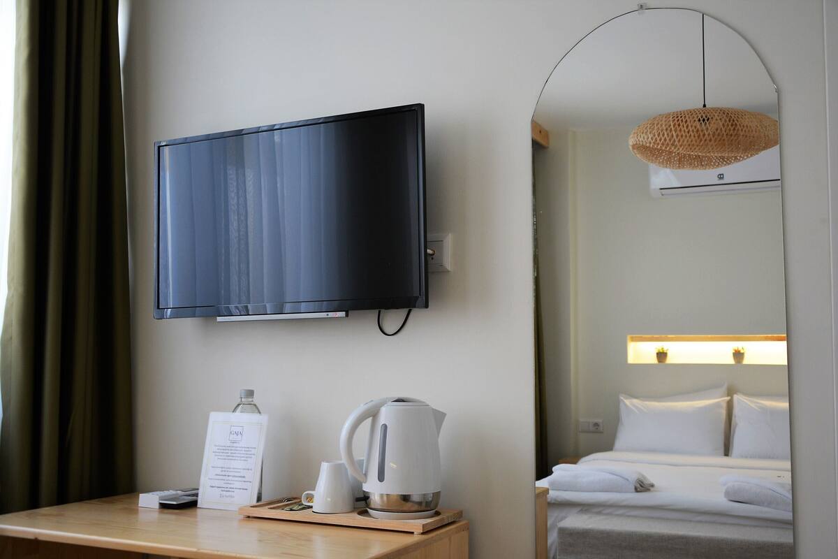 A hotel room with a flat-screen TV mounted on the wall, a wooden nightstand with a kettle, a sign, and a bottle.