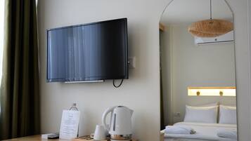 A hotel room with a flat-screen TV mounted on the wall, a wooden nightstand with a kettle, a sign, and a bottle.