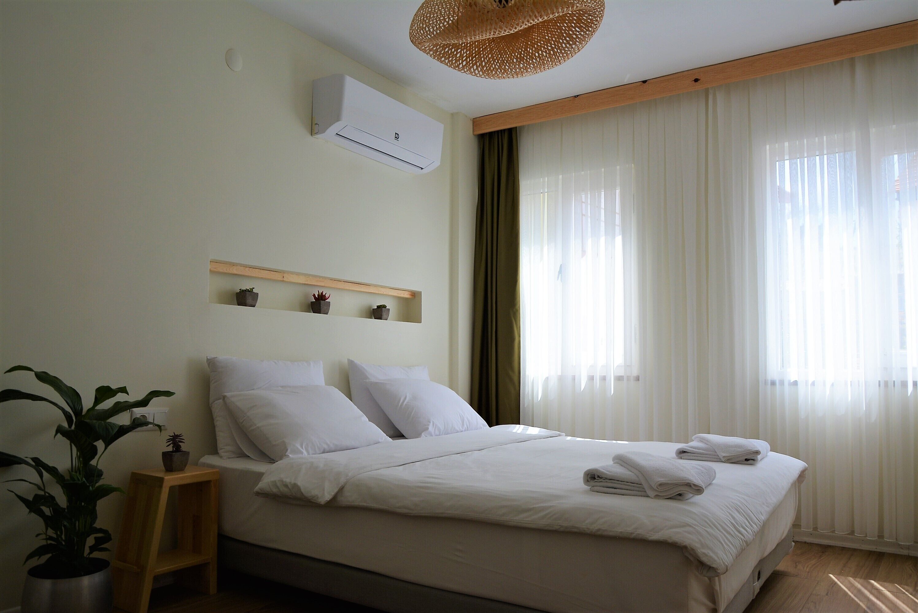 A neatly arranged bedroom with a bed, a bedside table with a plant, a wall-mounted air conditioner, and a window with sheer curtains.