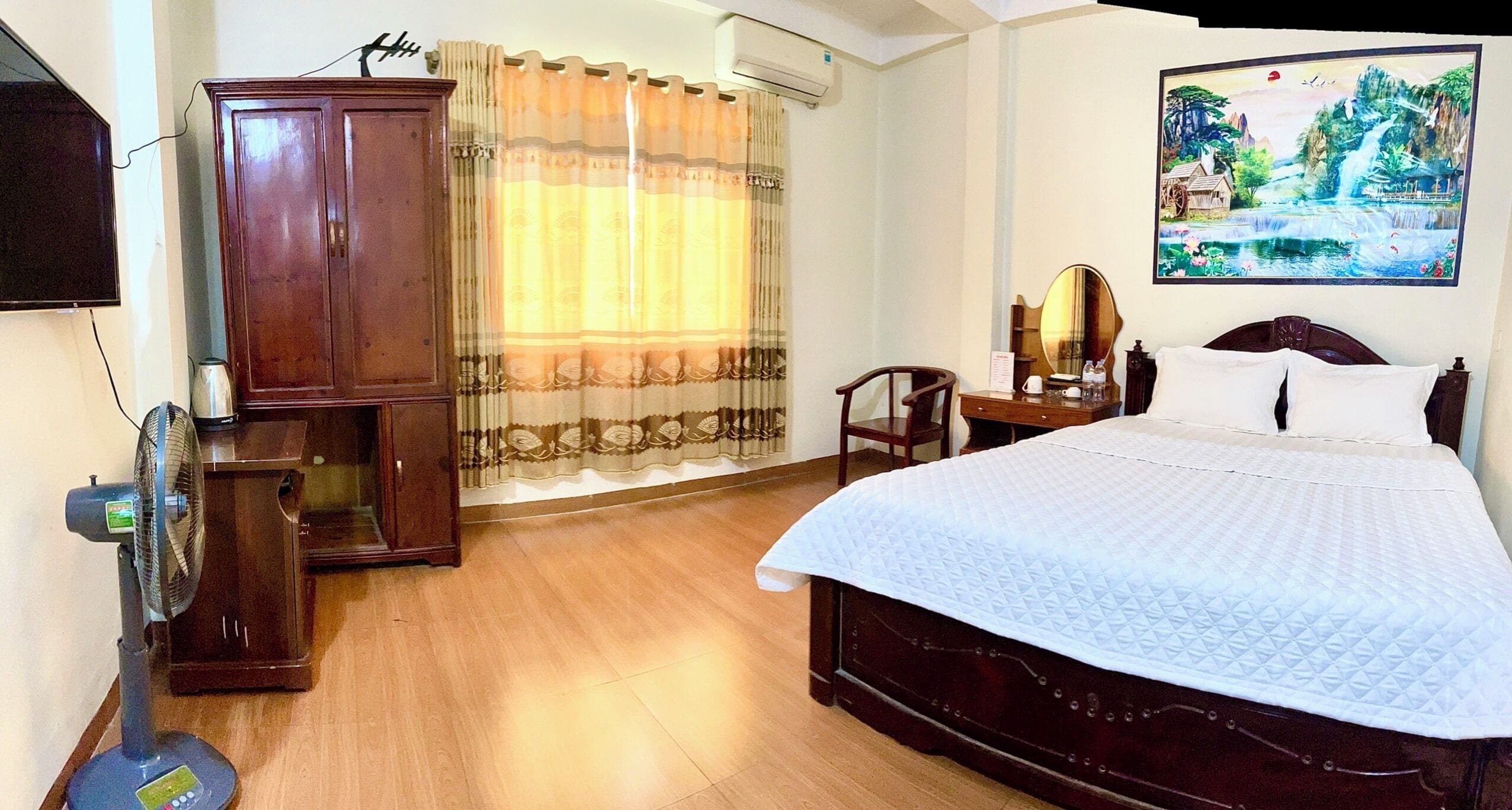 A hotel room with a bed, a desk, a chair, a wardrobe, and a painting on the wall.
