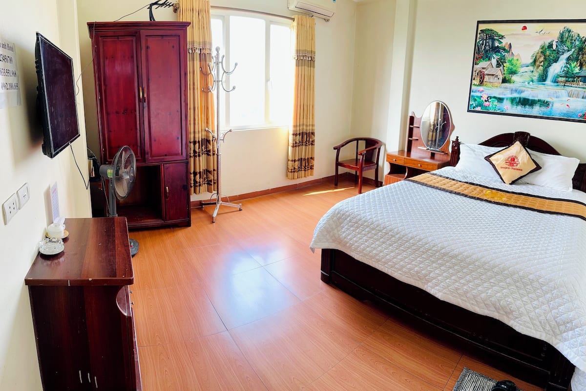 A hotel room with a bed, a wooden wardrobe, a desk with a chair, a TV, and a painting on the wall.