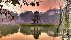 Um lago tranquilo com um pavilhão tradicional no centro, cercado por árvores e edifícios ao fundo.