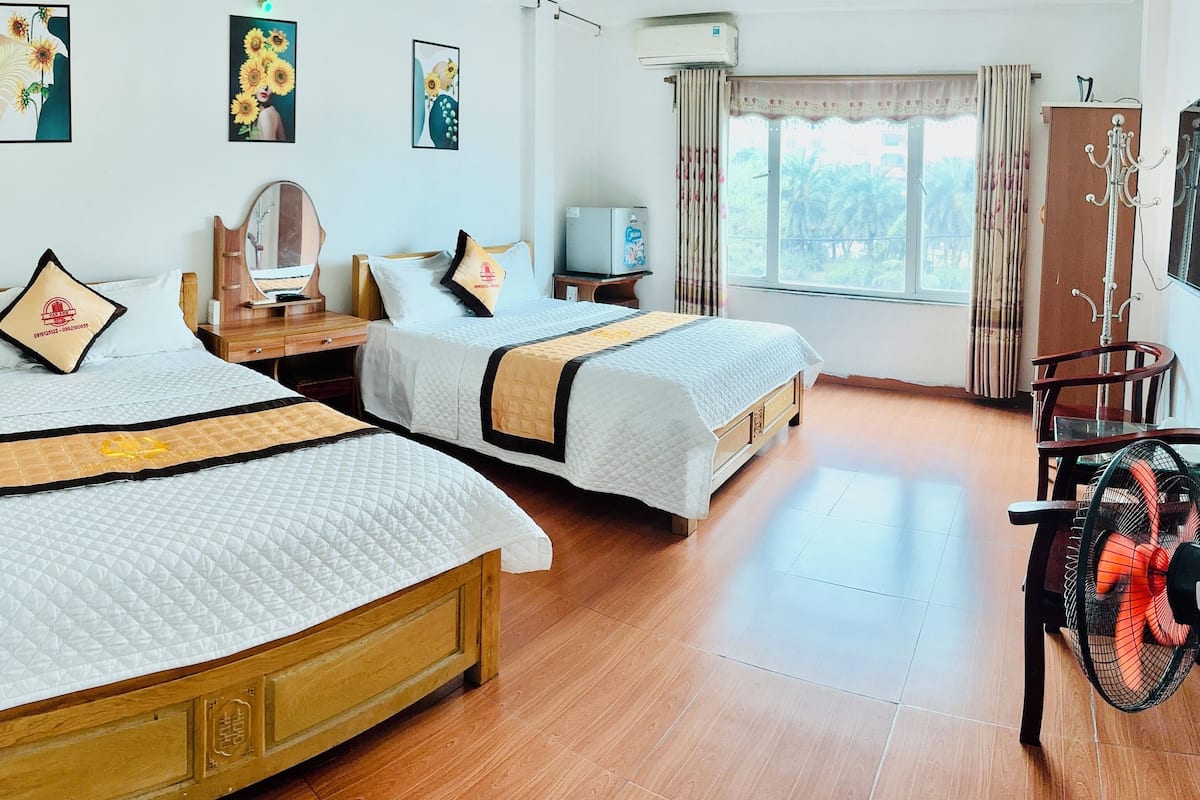 A hotel room with two beds, a wooden dresser, a mirror, a small table, a fan, and a window with curtains.