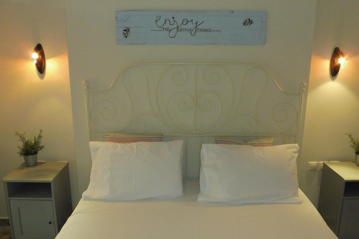 A bed with white linens, two pillows, a decorative headboard, bedside tables with plants, and wall sconces.
