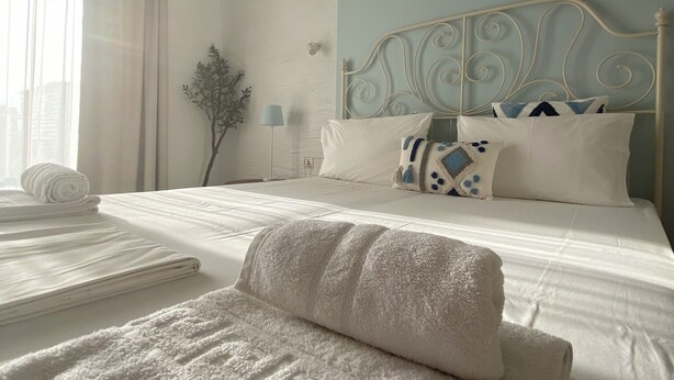 Una cama bien hecha con sábanas blancas y un cojín decorativo, una lámpara de noche y un aplique de pared.