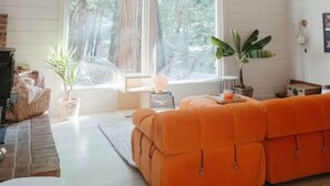 A spacious living room with a high ceiling and large windows, a bright orange sofa, and a fireplace. - Nevada City Cabin Pups and Long Stays Welcome (Nevada City)