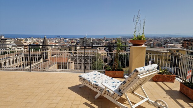 Eine Liege auf einer gefliesten Terrasse mit Blick auf die Stadt, einem Geländer und Pflanzkübeln.