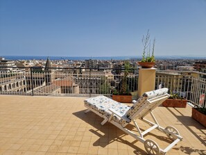 Eine Liege auf einer gefliesten Terrasse mit Blick auf die Stadt, einem Geländer und Pflanzkübeln.
