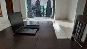 Un ordinateur portable est posé sur une table en bois dans une pièce dont le sol est carrelé, et d’où l’on aperçoit, par la fenêtre, un espace clos à l’extérieur.