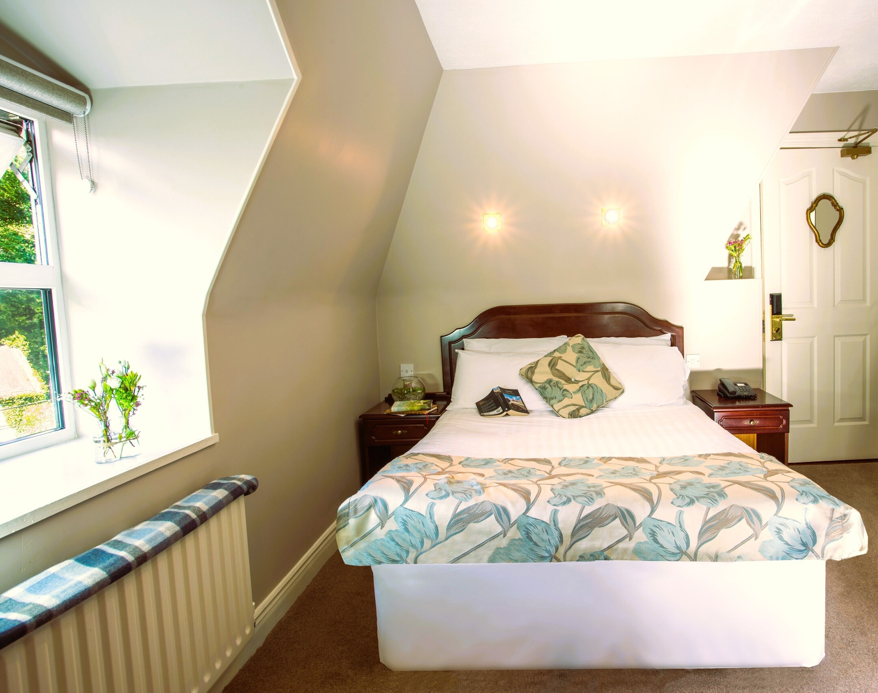 A bedroom with a large bed, a nightstand with a phone, a bedside table with a vase of flowers, and a window with a view of greenery.