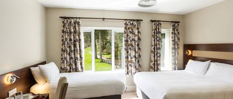 A hotel room with two beds, a desk with a chair, a window with curtains, and a view of greenery outside.