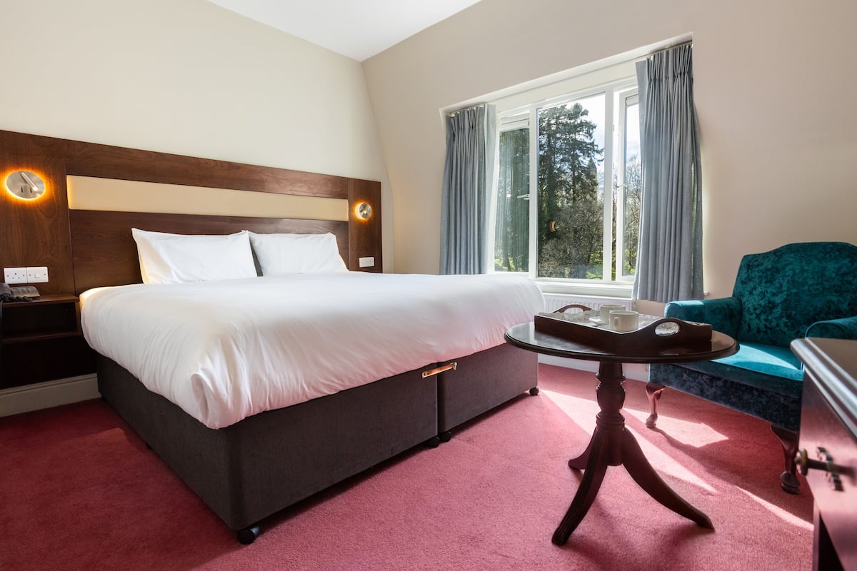 Een hotelkamer met een groot bed, een ronde tafel met dienblad, een blauwe fauteuil en een raam met gordijnen.