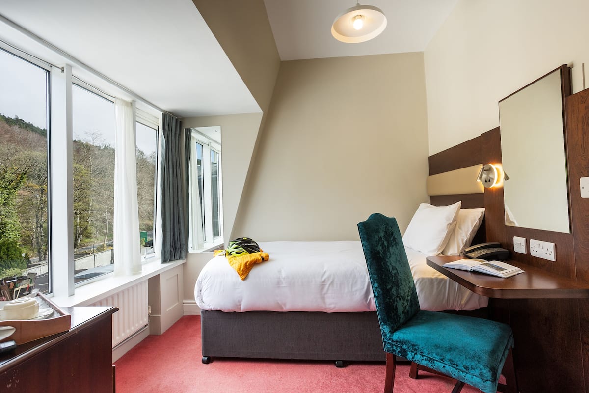 Een hotelkamer met een bed, een bureau, een stoel en een groot raam met uitzicht op bomen.