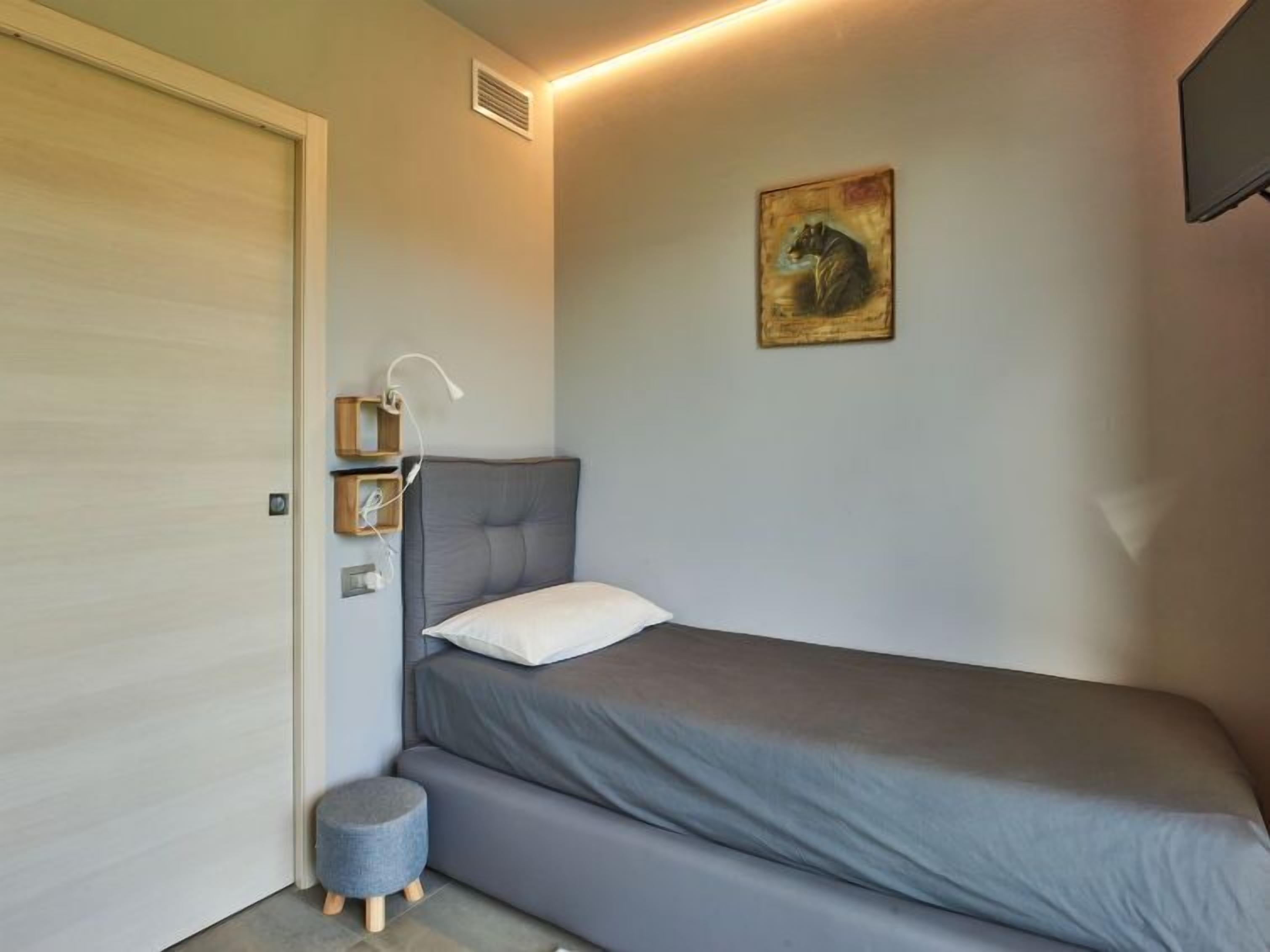 Een kleine, keurig opgemaakte slaapkamer met een enkel bed, een nachtkastje met een lamp, een klein krukje en een schilderij aan de muur.