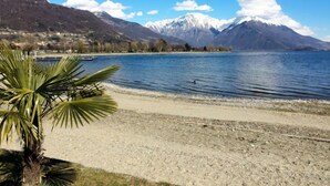 Um lago com montanhas ao fundo, uma praia de areia e uma palmeira em primeiro plano.