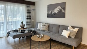 Una sala de estar moderna con un sofá gris, una mesa de centro redonda de madera y un cuadro de un paisaje montañoso en la pared.