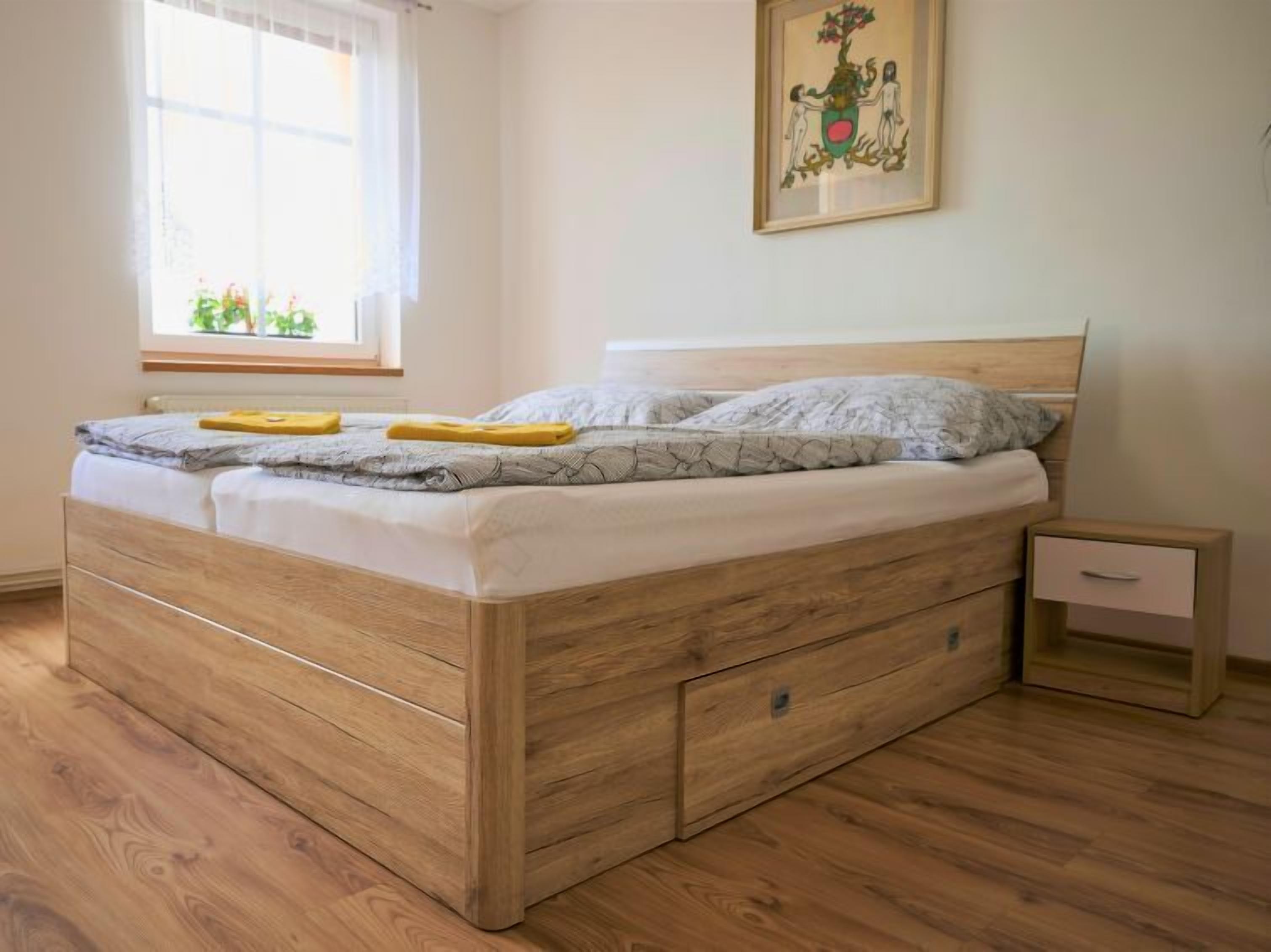 Cama doble con cabecera de madera y cajones, mesita de noche con un cajón y una ventana con cortinas.