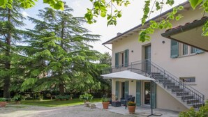 Un bâtiment à deux étages doté d’un patio couvert, d’un escalier et d’un jardin bien entretenu avec de grands arbres et des plantes en pot.