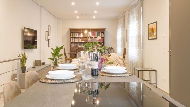Un coin repas avec une longue table dressée pour quatre personnes, entourée de chaises. On aperçoit, au fond, une bibliothèque remplie de livres.