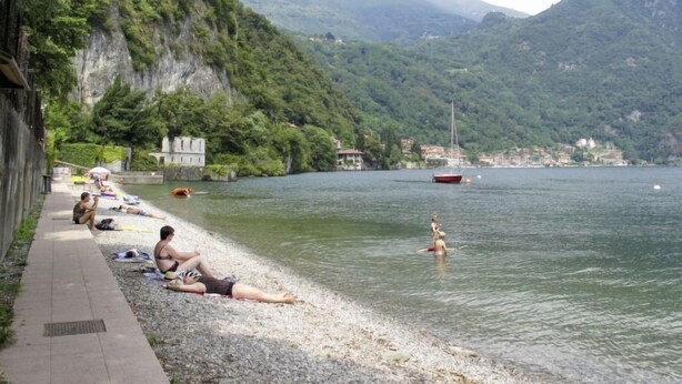 Une plage au bord d’un lac où des gens se prélassent au soleil et se baignent, le tout entouré de montagnes et avec une petite ville en arrière-plan.