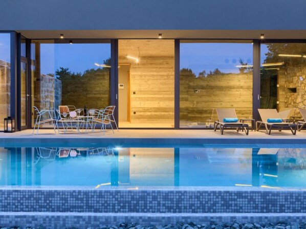 Un área de piscina moderna al aire libre con sillones reclinables y un porche con paredes de vidrio.