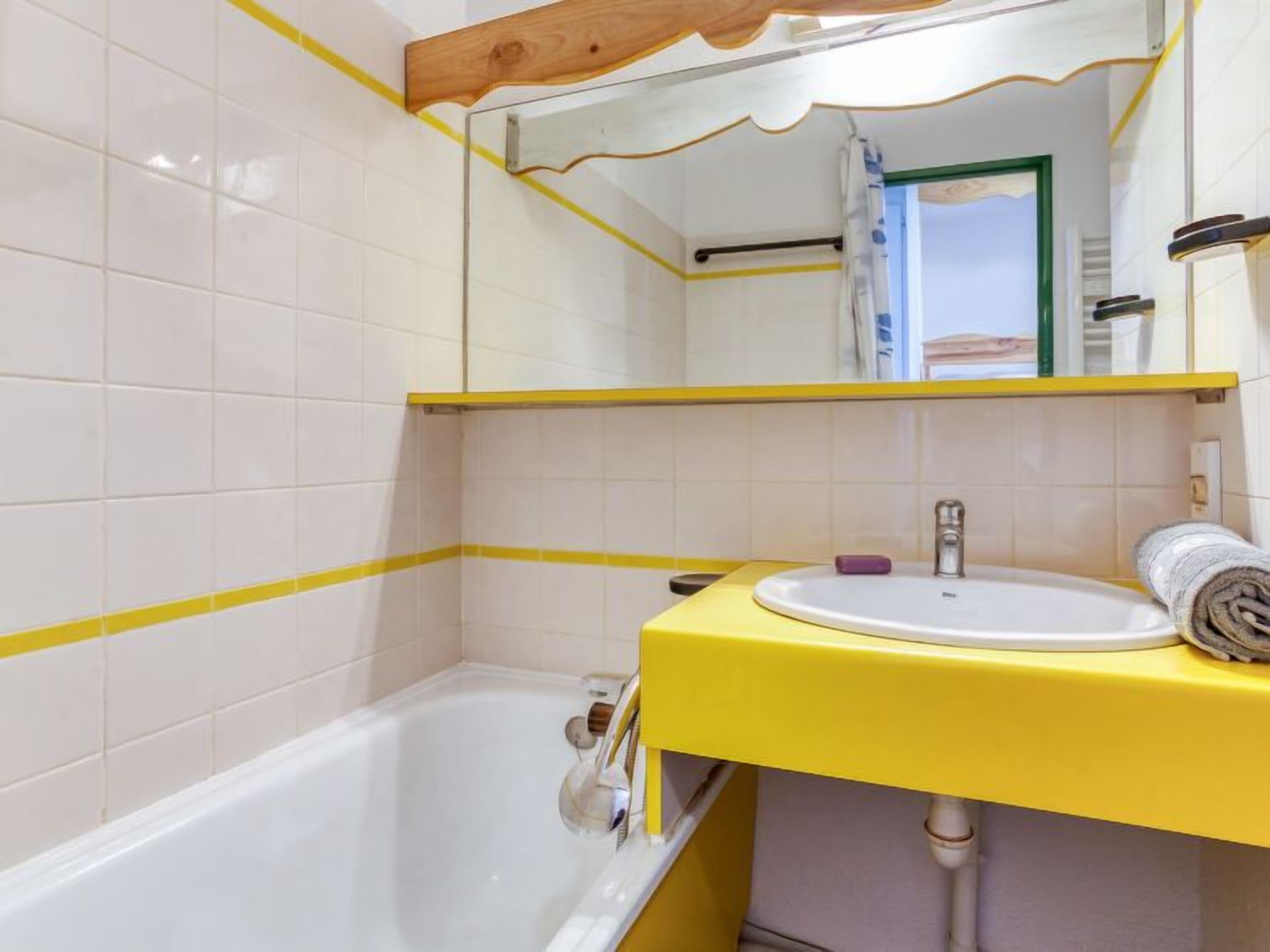 Ein Badezimmer mit weißen Fliesen, einem gelben Waschtisch, einer weißen Badewanne und einem Spiegel mit Holzrahmen.