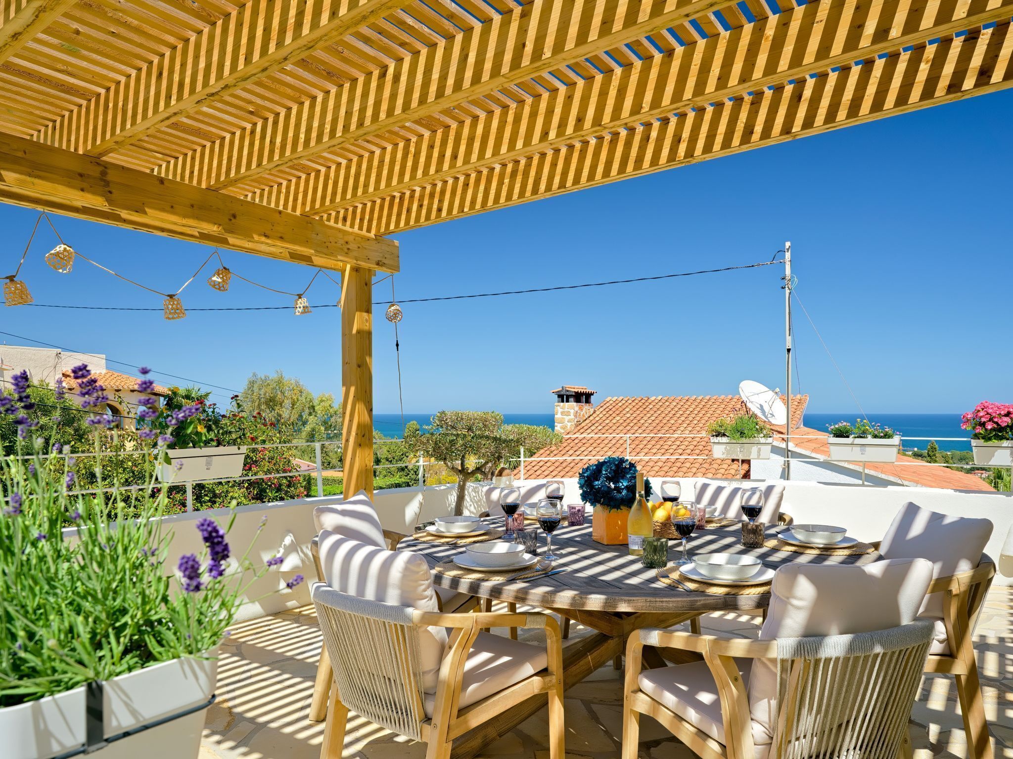 Eine Terrasse im Freien mit einer Holz-Pergola, einem runden Esstisch, Korbstühlen und Blick auf das Meer.