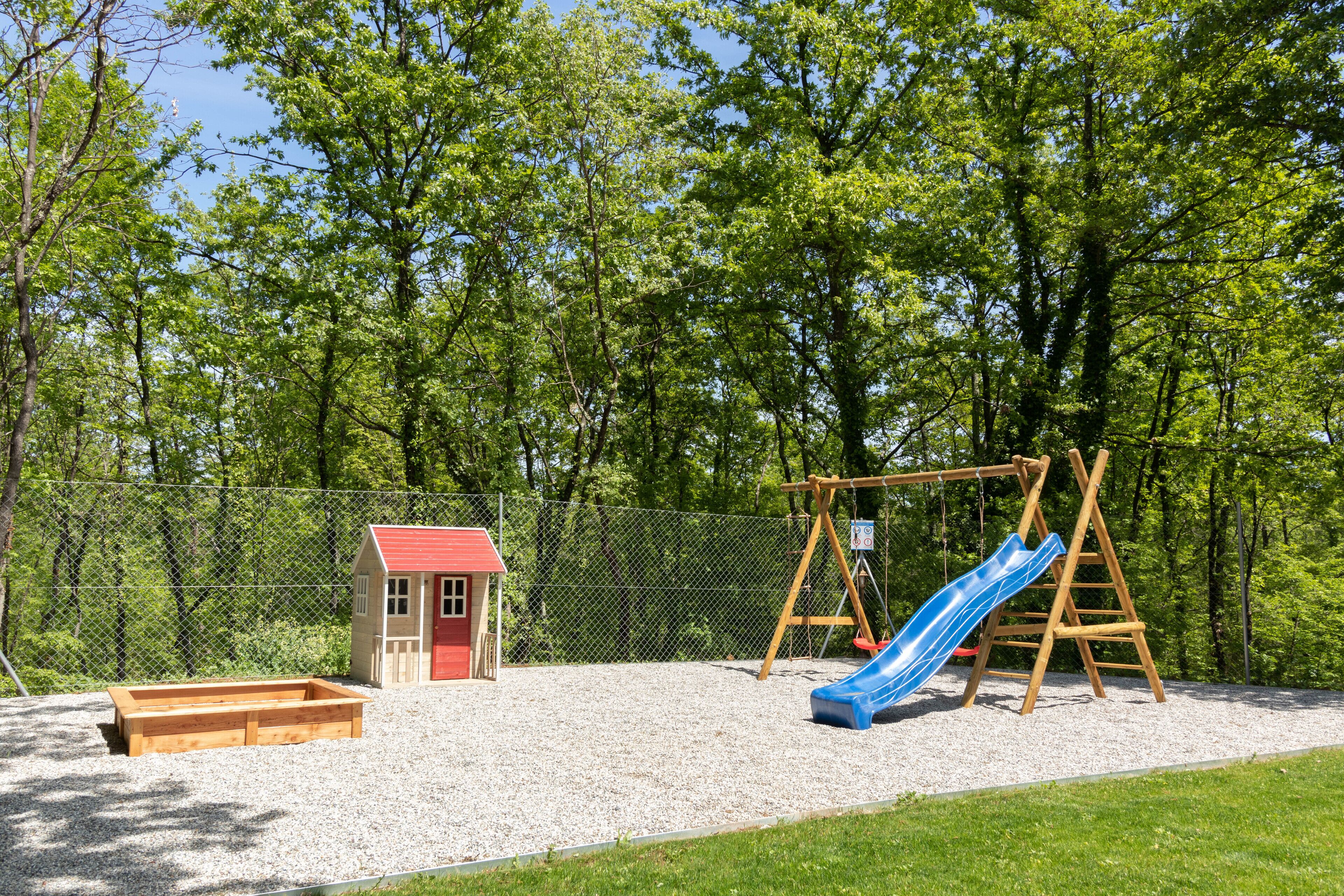 Une aire de jeux avec une balançoire en bois et un toboggan bleu, entourée d’une clôture et d’arbres.