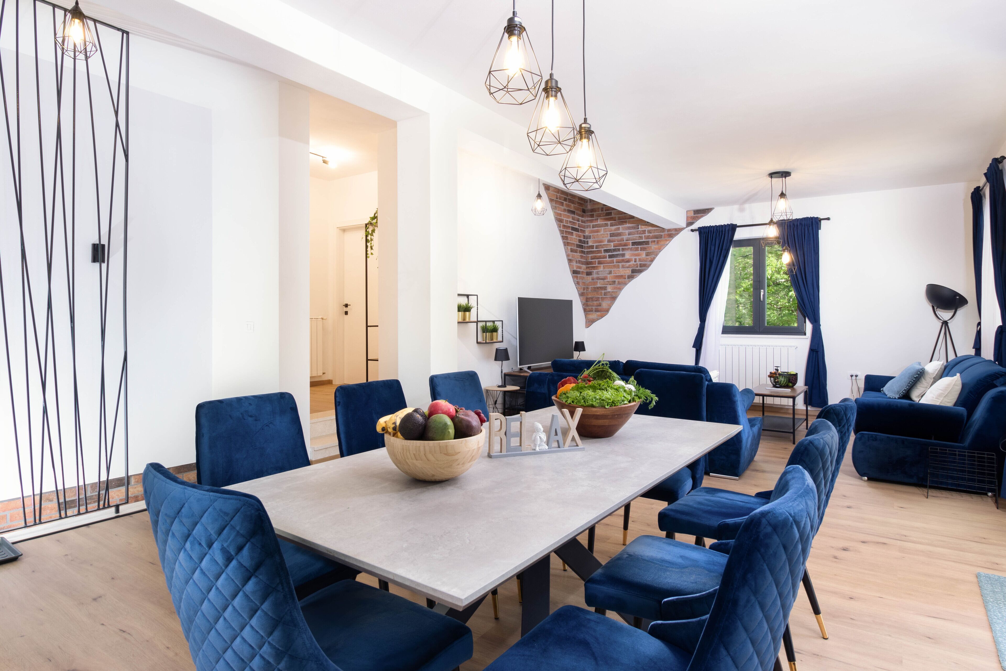 A modern dining area with a long table, blue upholstered chairs, and a bowl of fruit.