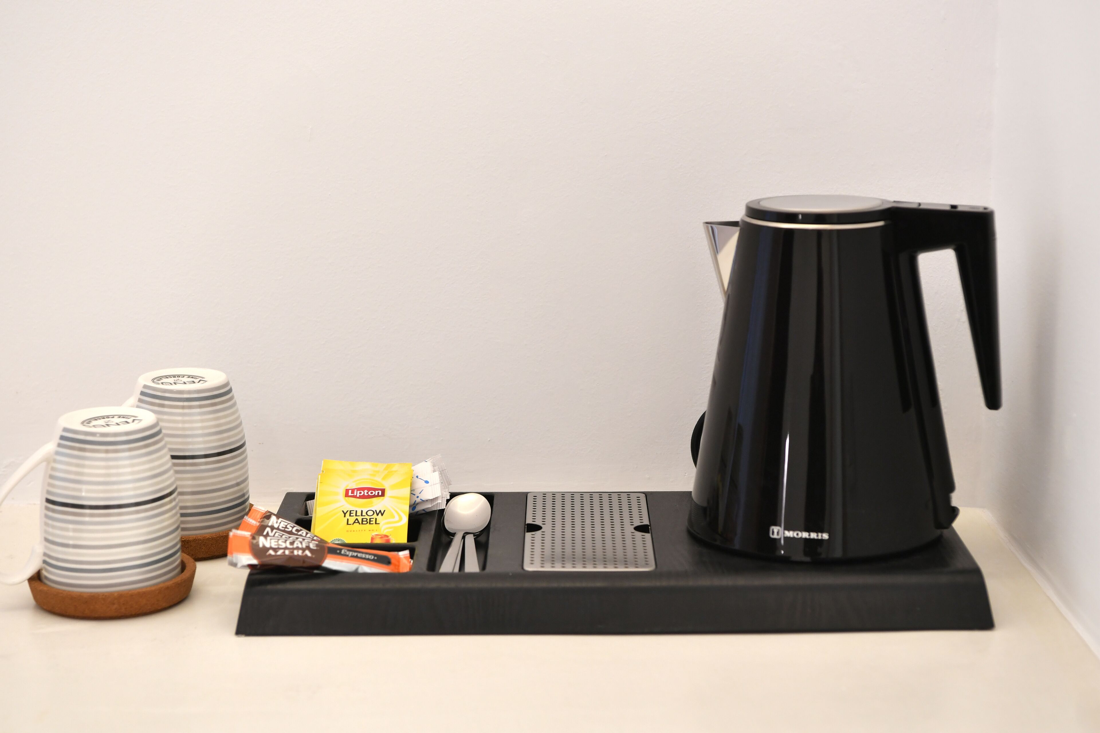A coffee maker, two stacked cups, a spoon, a packet of Lipton tea, and a packet of Nescafe coffee on a black mat.