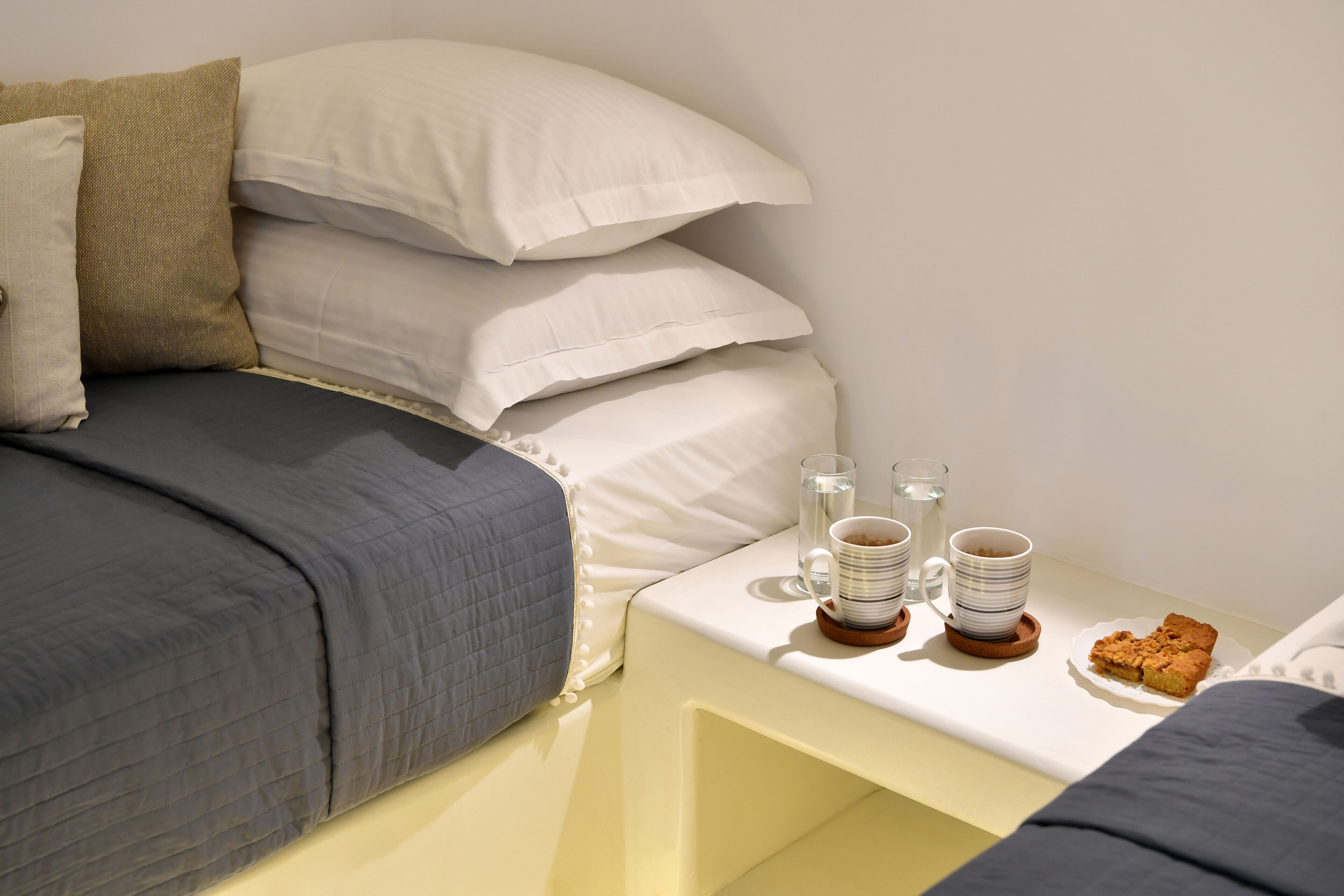 A neatly made bed with a gray and white duvet, a beige pillow, and a white pillow. There is a bedside table with a white tray holding a mug, a glass, and a plate with food.