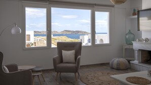 Ein Zimmer mit großem Fenster und Blick auf eine Küstenlandschaft, ein bequemer Sessel, ein kleiner runder Tisch und ein dekorativer Teppich.