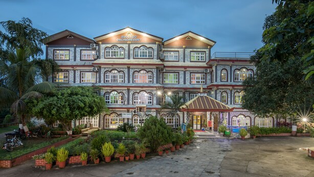 Un grand bâtiment à plusieurs étages, sur lequel on peut lire l’inscription « Avocado Orchid Resort ». Il possède une entrée centrale avec un passage couvert, entouré de plantes en pot et d’une cour dallée.
