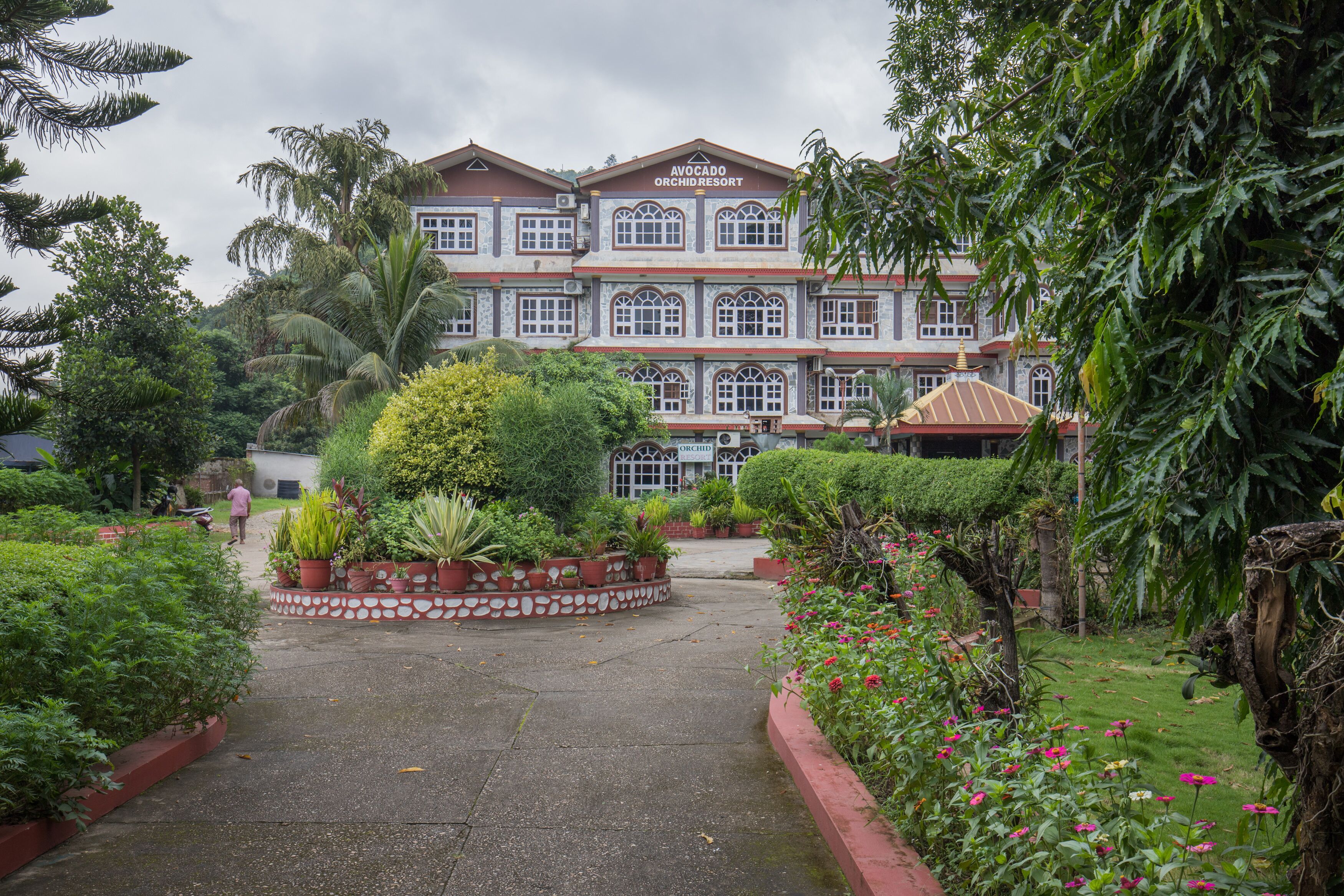 A large building with a sign reading "Avocado Orchid Resort." It features a central courtyard with potted plants and a well-maintained lawn.