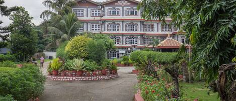 A large building with a sign reading "Avocado Orchid Resort." It features a central courtyard with potted plants and a well-maintained lawn.