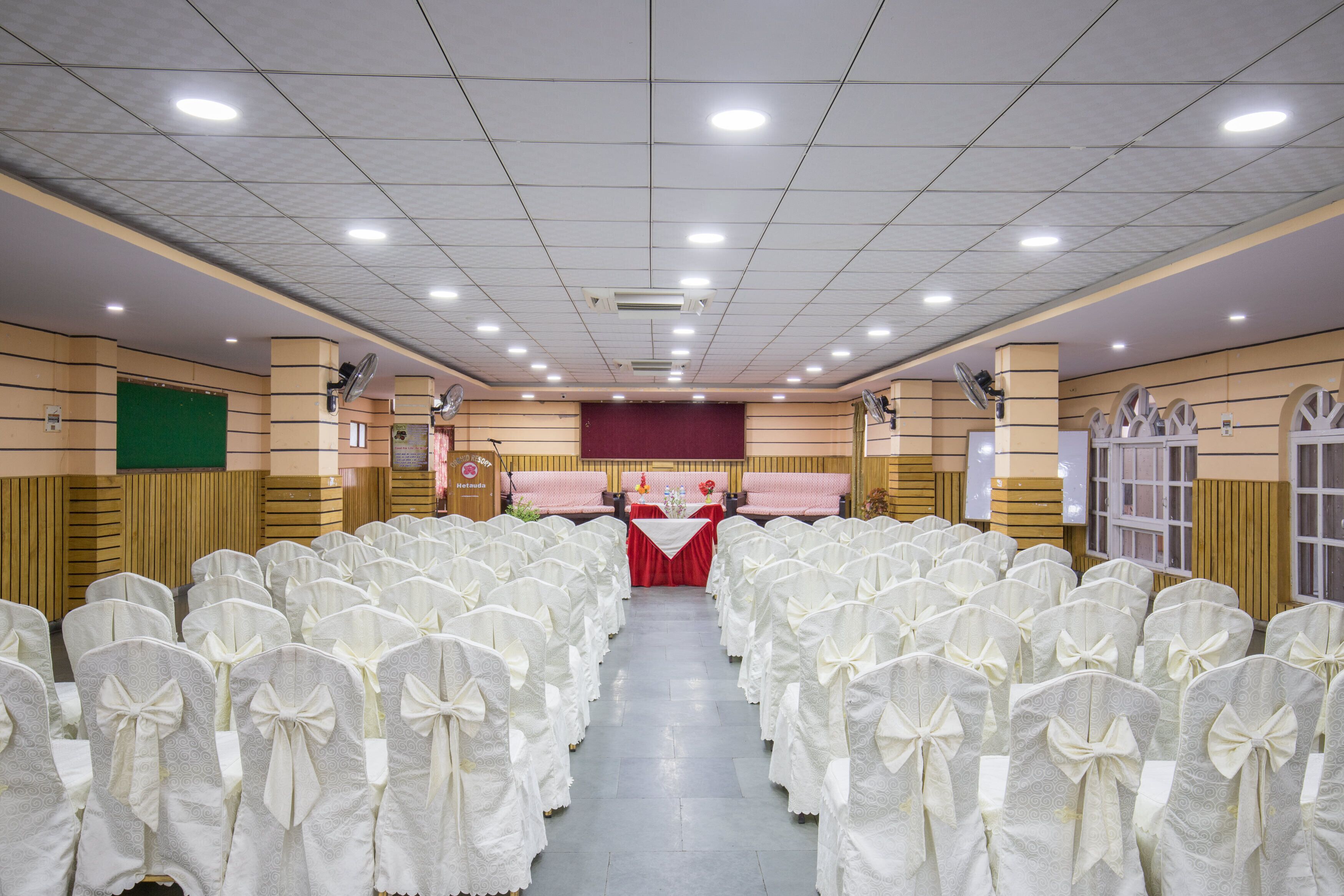 A banquet hall with rows of white chairs with bows, a stage with a red backdrop, and a ceiling with recessed lighting.