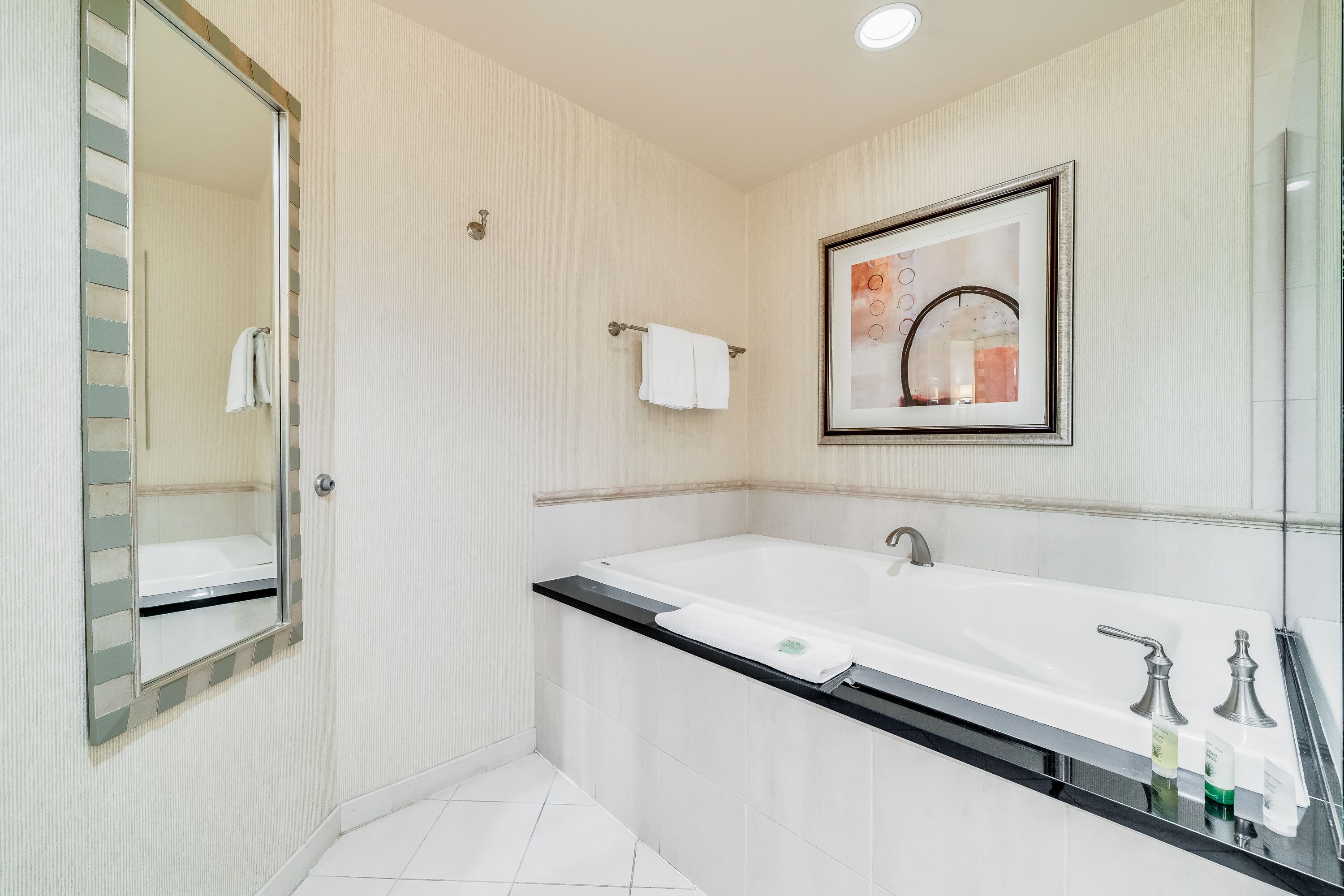 A modern bathroom with a large bathtub, a mirror, and a framed artwork on the wall.