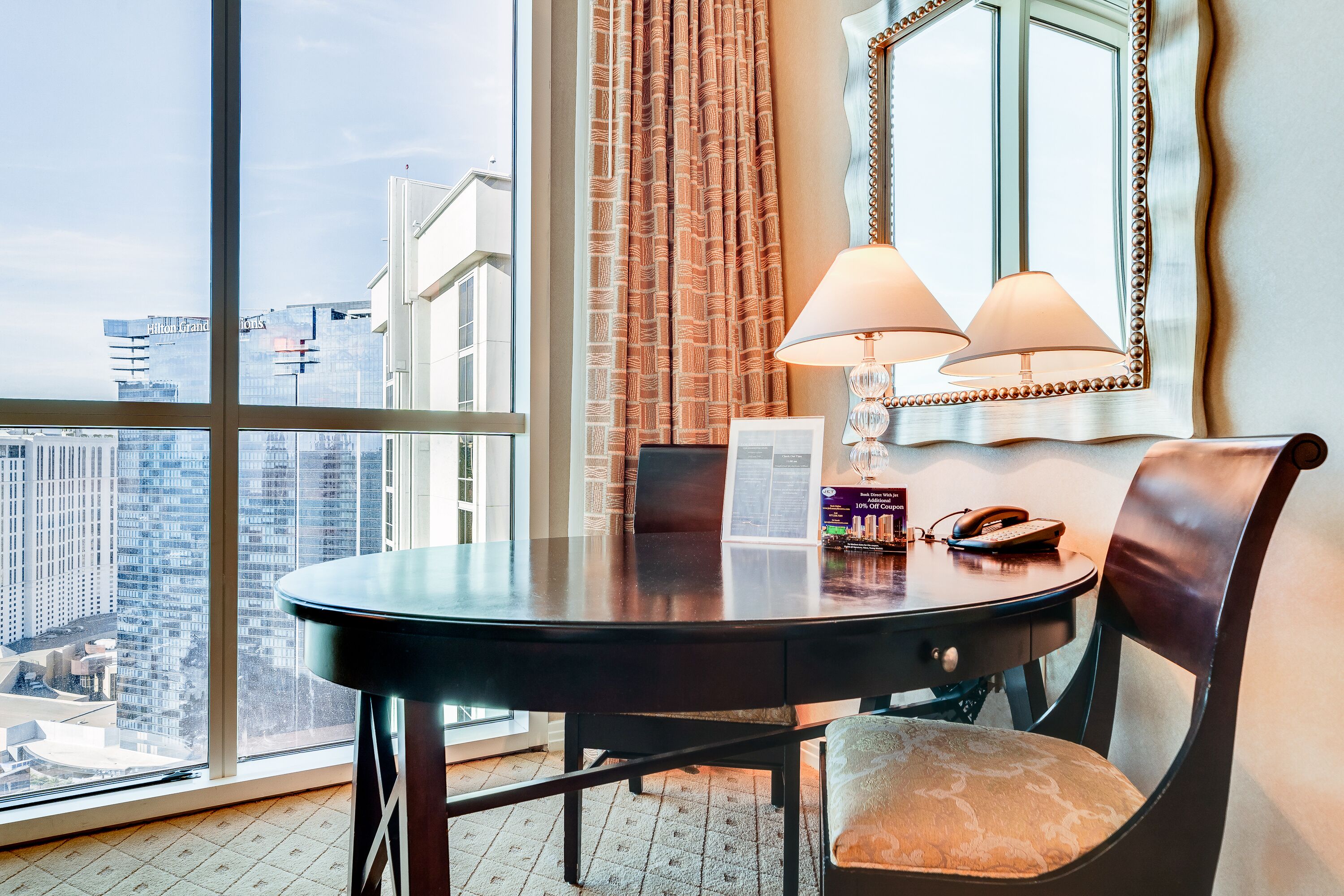 A hotel room with a large window, a round table with a chair, a lamp, and a telephone.